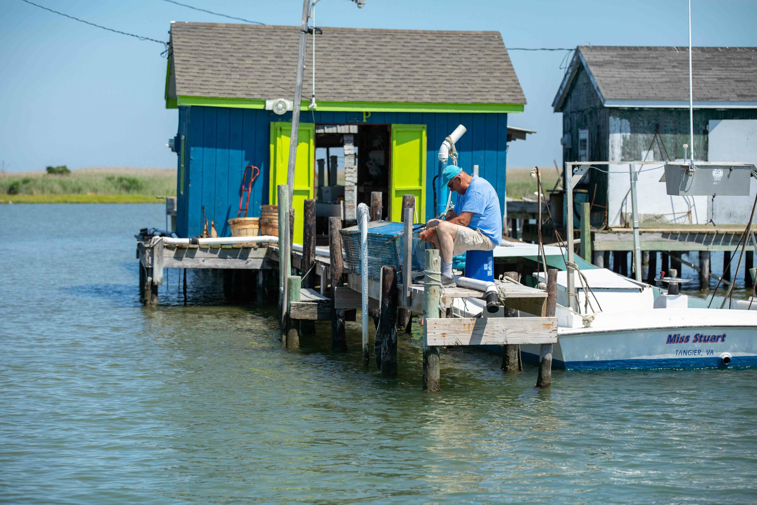 Man sits on dock in front of his blue crab shanty | VPM COM